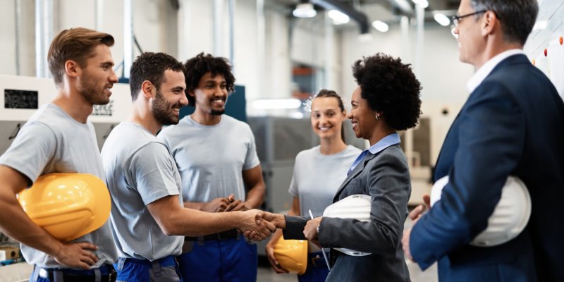 Como conquistar um bom relacionamento interpessoal no trabalho Uma mulher negra de terno aperta a mão de um trabalhador da indústria