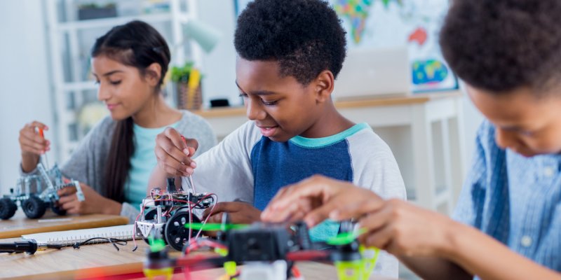 Tecnologia E Educação: O Aprendizado Através Da Robótica A imagem mostra três crianças sentadas em suas classes em uma sala de aula. Cada uma está mexendo em um robô de lego.