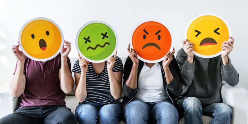 Quatro pessoas estão sentadas e seguram placas de diferentes emojis na frente dos rostos.
