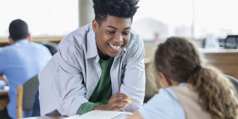 Vê-se um menino negro de camiseta em uma sala de aula, apoiado na mesa de uma colega e conversando com ela sorrindo.